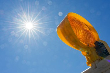 Sarı bir inşaat alanı uyarı lambası düşük güneşi yakalar ve her yöne ince ışınlar gönderir. Parlak alev, berrak ve sakin bir gökyüzüne karşı kurulmuş basit yol çalışmalarına ışıltı katıyor..