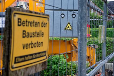 Aynı sarı tabela çitin arkasında asılıyken yolun ilerleyişi devam ediyor. Elinde kova olan sarı bir kazıcı toprak yığınları ve işaretlerin arasında duruyor. Etkin bir inşaat bölgesinde makine ve bariyerler