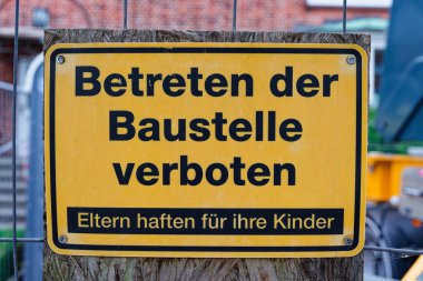 İnce bir çubuk çitin üzerinde Betreten der Baustelle verboten yazan sarı bir metal tabela vardır. Çizginin altında Eltern, Kinder 'ın ortaya çıkacağı yer var. Tabak küçük bir tahtaya vidalanmış..
