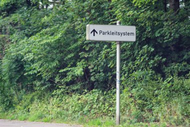 Metal bir yol kenarı direğinde beyaz bir plaka var. Önünde bir ok var. Sahne iki şeritli sessiz bir yol gösteriyor, temiz kenar, alçak çalılar ve gün ışığı yeşillik boyunca..