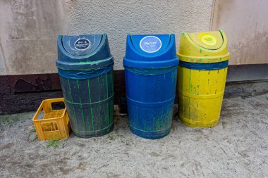 Üç ahşap çöp kutusu beton bir avluda duruyor. Renkli sarı, mavi ve koyu mavi. Etiketlerde biyomuell, kağıt ve gelbersack yazıyor. Sarı bir sandık yanlarında duruyor, dönmeye hazır..