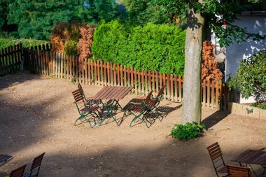 Konforlu teras, her biri düzgün bahçe yataklarının yanında sandalyeli katlanabilir masalar barındırır. Düşük ahşap çit çerçeve çalılar ve düzenli bir yol, birkaç kuru yaprak hakkında yalan söyler ama ortama özen gösterilir ve sakin gelir..