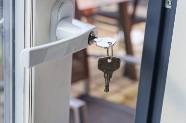 Kapı terasa hafifçe açık duruyor. Dışarıda küçük masalar bir klinik ya da kafe gibi görünüyor. Kilitte bırakılmış bir anahtar erişimi kontrol etmeye ve açma limitini korumaya yardımcı olur.