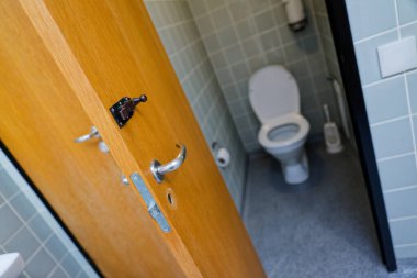 Küçük açık tuvalet manzarası ahşap kapının geniş açıyla sallandığını gösteriyor. İçinde parlak beyaz seramik bir tuvalet var gri fayanslara karşı basit bir cıvatayla ve yumuşak ışık altında..