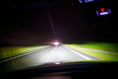 Ağır bir gece gökyüzünün altındaki yeşil tarlalar arasında karanlık bir otoyol kesiliyor. İleride ufukta parlak bir parıltı beliriyor. Bu sırada geçen arabalar ışığın yumuşak izlerini boyuyor. Sahne boyunca bulanık..