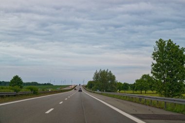 Geniş otoban, iki şeritte de sabit trafiği taşır. Kenarlarında metal korkuluklar ve uzun ağaçlar yol boyunca uzanır. Arabalar ise temiz bir gökyüzünün altında güvenli bir şekilde ilerler..
