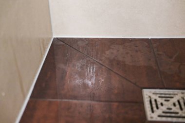 Yüzeyinde temiz su parlarken beyaz duvarları ve ıslak kahverengi zemini olan fayanslı bir duş kabini çerçeveler ve parlak banyoda taze buhar kalır..