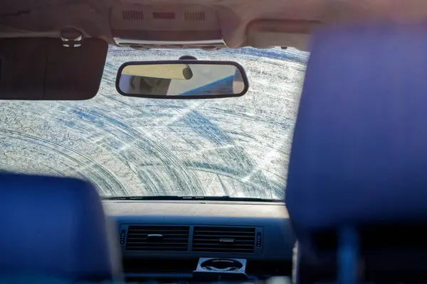 The front windshield is coated with dirt and smeared streaks, visibility is zero, light diffuses across the grime and haze, wipers have left uneven traces, the car is unsafe to drive until cleaned