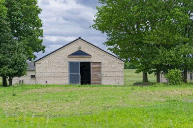 Yüksek bir tarım salonu iki geniş sürgülü kapısı ve penceresi olmayan açık bir zemin üzerinde duruyor. Ağaçlar gün ışığında sessiz yeşil bir zemin oluştururken, basit metal kaplı bina kısa bir çimenliğe bakıyor..