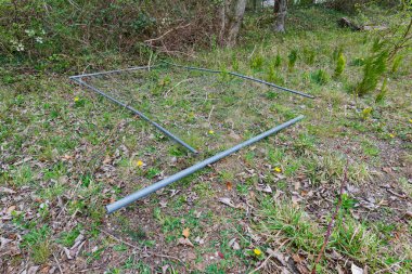 Four thin gray pipes lie side by side on grass near the edge of a wood. The tubes look new and clean, likely waiting for a small project or repair, while quiet trees stand beyond under a soft sky.