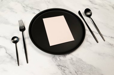 Siyah çatal bıçak takımı ve siyah plaka beyaz mermer arka planda. Masa ayarı menüsü, boş kağıt düzlüğü. Çatal, bıçak, kaşık, tatlı kaşığı, tabak ve boş. Boşluğu kopyala.