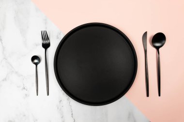Siyah çatal bıçak takımı ve siyah plaka beyaz mermer ve pembe arka planda. Yaratıcı masa düzleştirici. Çatal, bıçak, kaşık, tatlı kaşığı ve tabak. Boşluğu kopyala.
