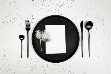 Noel kompozisyonu, siyah tabak ve çatal bıçak takımı, beyaz kart, köknar ağacı dalı, beyaz arka planda gümüş yıldız konfetisi. Noel, yeni yıl, kış konsepti. Üst görünüm, düz görünüm, kopyalama alanı. 