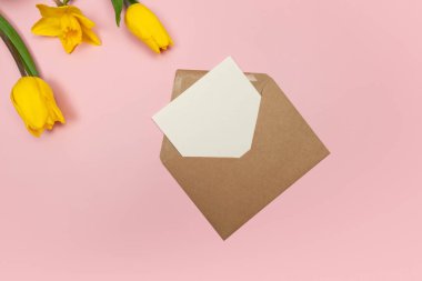 Sarı lalelerin ve pembe arka plandaki nergislerin üst görüntüsü. Kraft zarfı, beyaz kart. Bahar renkli kompozisyon, düz yatış, kopyalama alanı.
