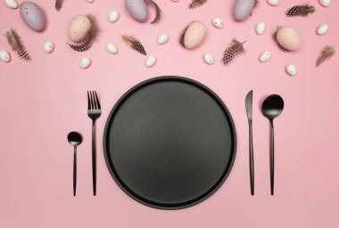 Paskalya yumurtaları, tüyler, siyah tabak ve pembe arka planda çatal bıçak takımı. Yaratıcı paskalya kompozisyonu, yay, düz yatış, kopyalama alanı.