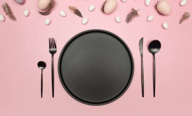 Paskalya yumurtaları, tüyler, siyah tabak ve pembe arka planda çatal bıçak takımı. Yaratıcı paskalya kompozisyonu, yay, düz yatış, kopyalama alanı.