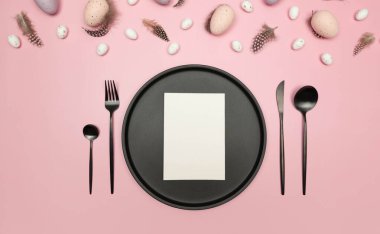 Paskalya yumurtaları, tüyler, beyaz kart, siyah tabak ve pembe arka planda çatal bıçak. Yaratıcı paskalya kompozisyonu, yay, düz yatış, kopyalama alanı.