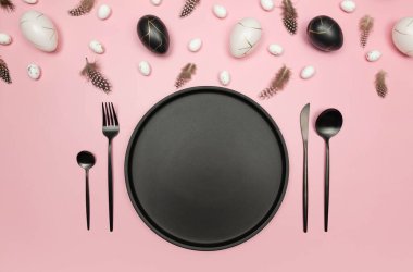 Paskalya yumurtalarının üst görüntüsü, pembe arka planda tüyler. Masa seti, siyah tabak ve çatal bıçak takımı, beyaz kart. Yaratıcı paskalya kompozisyonu, yay, düz yatış, kopyalama alanı.