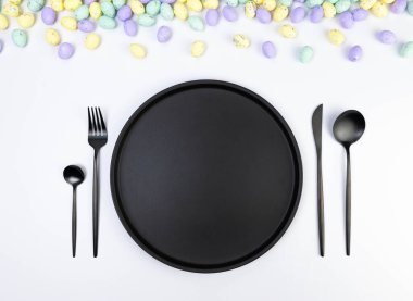 Beyaz arka planda renkli paskalya yumurtalarının üst görüntüsü. Masa takımı, siyah tabak ve çatal bıçak takımı. Yaratıcı paskalya kompozisyonu, yay, kopyalama alanı, düz konum.