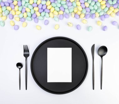 Beyaz arka planda renkli paskalya yumurtalarının üst görüntüsü. Masa takımı, beyaz kart, siyah tabak ve çatal bıçak takımı. Yaratıcı paskalya kompozisyonu, yay, kopyalama alanı, düz konum.