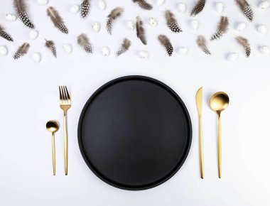 Kahverengi tüylerin üst görüntüsü, beyaz arka planda beyaz paskalya yumurtaları. Masa seti, siyah tabak ve altın çatal bıçak takımı. Yaratıcı paskalya kompozisyonu, yay, kopyalama alanı, düz konum. 