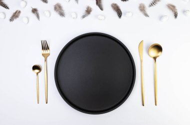 Kahverengi tüylerin üst görüntüsü, beyaz arka planda beyaz paskalya yumurtaları. Masa seti, siyah tabak ve altın çatal bıçak takımı. Yaratıcı paskalya kompozisyonu, yay, kopyalama alanı, düz konum. 