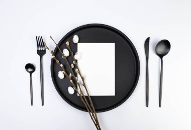 Top view of black plate and cutlery, white card, pussy willow branches, white easter eggs on white background. Spring easter composition, copy space, flat lay.