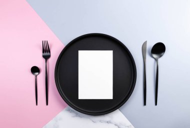 Siyah tabakların ve çatal bıçakların üst görüntüsü, renkli arka planda beyaz kart. Masa seti, mavi, pembe ve mermer arka plan. Pastel renkli duvar kağıdı, mermer doku, boşluğu kopyala.