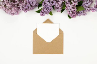 Beyaz arka planda mor leylak çiçeklerinin üst görüntüsü. Kahverengi kraft zarf, beyaz kart, posta. Yay duvar kağıdı, düz yatak, kopyalama alanı.