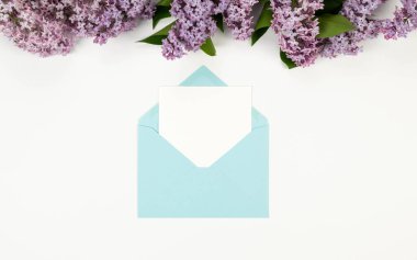 Beyaz arka planda mor leylak çiçeklerinin üst görüntüsü. Mavi zarf, beyaz kart, posta. Yay duvar kağıdı, düz yatak, kopyalama alanı.