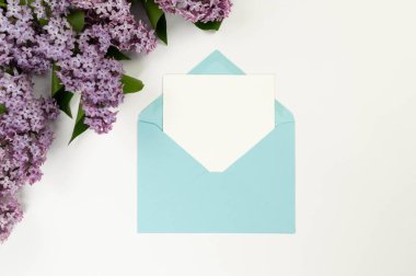 Beyaz arka planda mor leylak çiçeklerinin üst görüntüsü. Mavi zarf, beyaz kart, posta. Yay duvar kağıdı, düz yatak, kopyalama alanı.