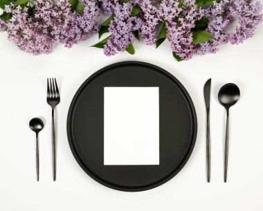 Beyaz arka planda mor leylak çiçeklerinin üst görüntüsü. Masa takımı, siyah tabak, siyah çatal bıçak takımı, beyaz kart. Yay duvar kağıdı, düz yatak, kopyalama alanı.