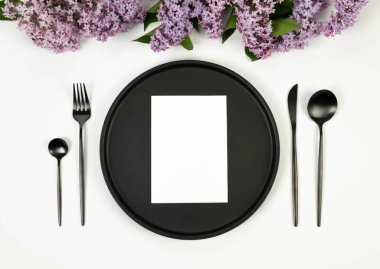 Beyaz arka planda mor leylak çiçeklerinin üst görüntüsü. Masa takımı, siyah tabak, siyah çatal bıçak takımı, beyaz kart. Yay duvar kağıdı, düz yatak, kopyalama alanı.