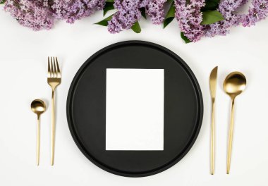 Beyaz arka planda mor leylak çiçeklerinin üst görüntüsü. Masa takımı, siyah tabak, altın çatal bıçak takımı ve beyaz kart. Yay duvar kağıdı, düz yatak, kopyalama alanı.
