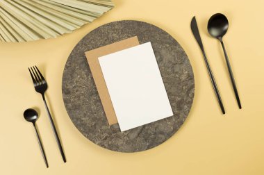 Kahverengi mermer plaka, siyah çatal bıçak takımı, sarı arka planda beyaz ve kahverengi kartlar. Kurumuş palmiye yaprağı. Yemek sunumu, taş tepsi. Tablo ayarı, kopyalama alanı, düz uzanma.