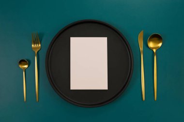 Siyah tabağın üst görüntüsü, altın çatal bıçak takımı ve koyu yeşil arka planda pembe kart. Yemek sunumu, siyah tepsi. Tablo ayarı, kopyalama alanı, düz uzanma.