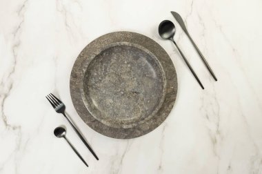 Kahverengi mermer tepsi ve tabağın üst görüntüsü, beyaz mermer arka planda siyah çatal bıçak takımı. Yemek sunumu, taş tepsi. Tablo ayarı, kopyalama alanı, düz uzanma.