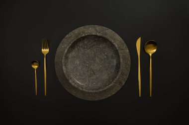 Kahverengi mermer tepsi ve tabağın üst görüntüsü, siyah arka planda altın çatal bıçak takımı. Yemek sunumu, taş tepsi. Tablo ayarı, kopyalama alanı, düz uzanma.