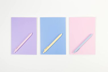 Beyaz arka planda pembe, mor ve mavi defterlerin üst görüntüsü. Pembe, sarı ve mavi kalemler, okul, ofis kağıdı. Düz yat, boşluğu kopyala.