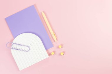 Mor defterin üst görüntüsü, sarı kalem, ataç, raptiyeler, pembe arka planda beyaz kemer tepsisi. Okul, ofis duvar kağıdı. Düz yat, boşluğu kopyala.