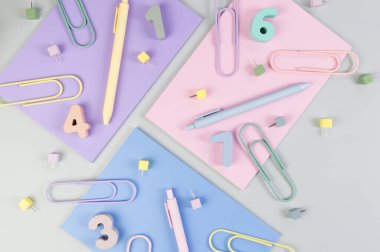 Gri arkaplanda pembe, mor ve mavi defterlerin üst görüntüsü. Renkli kalemler, ataçlar, numaralar ve iğneler. Okul, ofis duvar kağıdı. Düz yat, boşluğu kopyala.