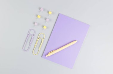 Gri arkaplandaki mor defterin üst görüntüsü. Ataç, kalem ve raptiye. Okul, ofis duvar kağıdı. Düz yat, boşluğu kopyala.