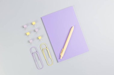 Gri arkaplandaki mor defterin üst görüntüsü. Ataç, kalem ve raptiye. Okul, ofis duvar kağıdı. Düz yat, boşluğu kopyala.