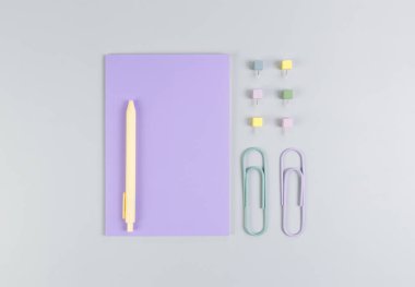 Gri arkaplandaki mor defterin üst görüntüsü. Ataç, kalem ve raptiye. Okul, ofis duvar kağıdı. Düz yat, boşluğu kopyala.