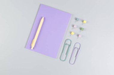 Gri arkaplandaki mor defterin üst görüntüsü. Ataç, kalem ve raptiye. Okul, ofis duvar kağıdı. Düz yat, boşluğu kopyala.