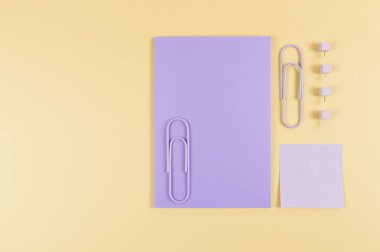 Sarı arkaplanda mor defterin üst görüntüsü. Mor yapışkan not, ataç ve raptiyeler. Okul, ofis duvar kağıdı. Düz yat, boşluğu kopyala.