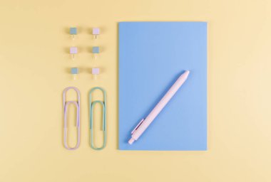 Sarı arkaplanda mavi defterin üst görüntüsü. Ataç, kalem ve raptiye. Okul, ofis duvar kağıdı. Düz yat, boşluğu kopyala.