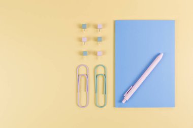 Sarı arkaplanda mavi defterin üst görüntüsü. Ataç, kalem ve raptiye. Okul, ofis duvar kağıdı. Düz yat, boşluğu kopyala.