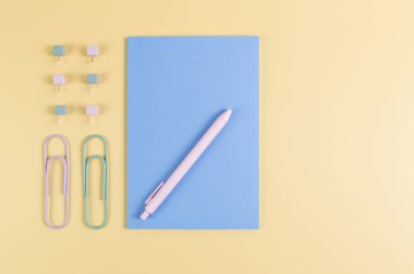 Sarı arkaplanda mavi defterin üst görüntüsü. Ataç, kalem ve raptiye. Okul, ofis duvar kağıdı. Düz yat, boşluğu kopyala.