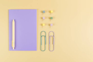 Sarı arkaplanda mor defterin üst görüntüsü. Ataç, kalem ve raptiye. Okul, ofis duvar kağıdı. Düz yat, boşluğu kopyala.